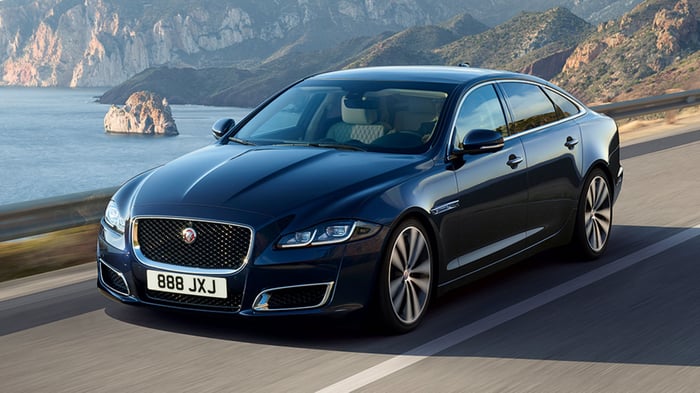 Dark-colored Jaguar on a road in front of a seaside landscape.