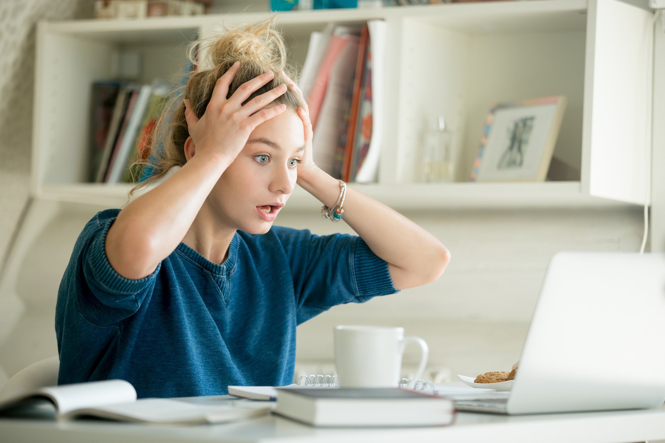 Woman at a table grabbing her head in surprise or disbelief while looking at her laptop