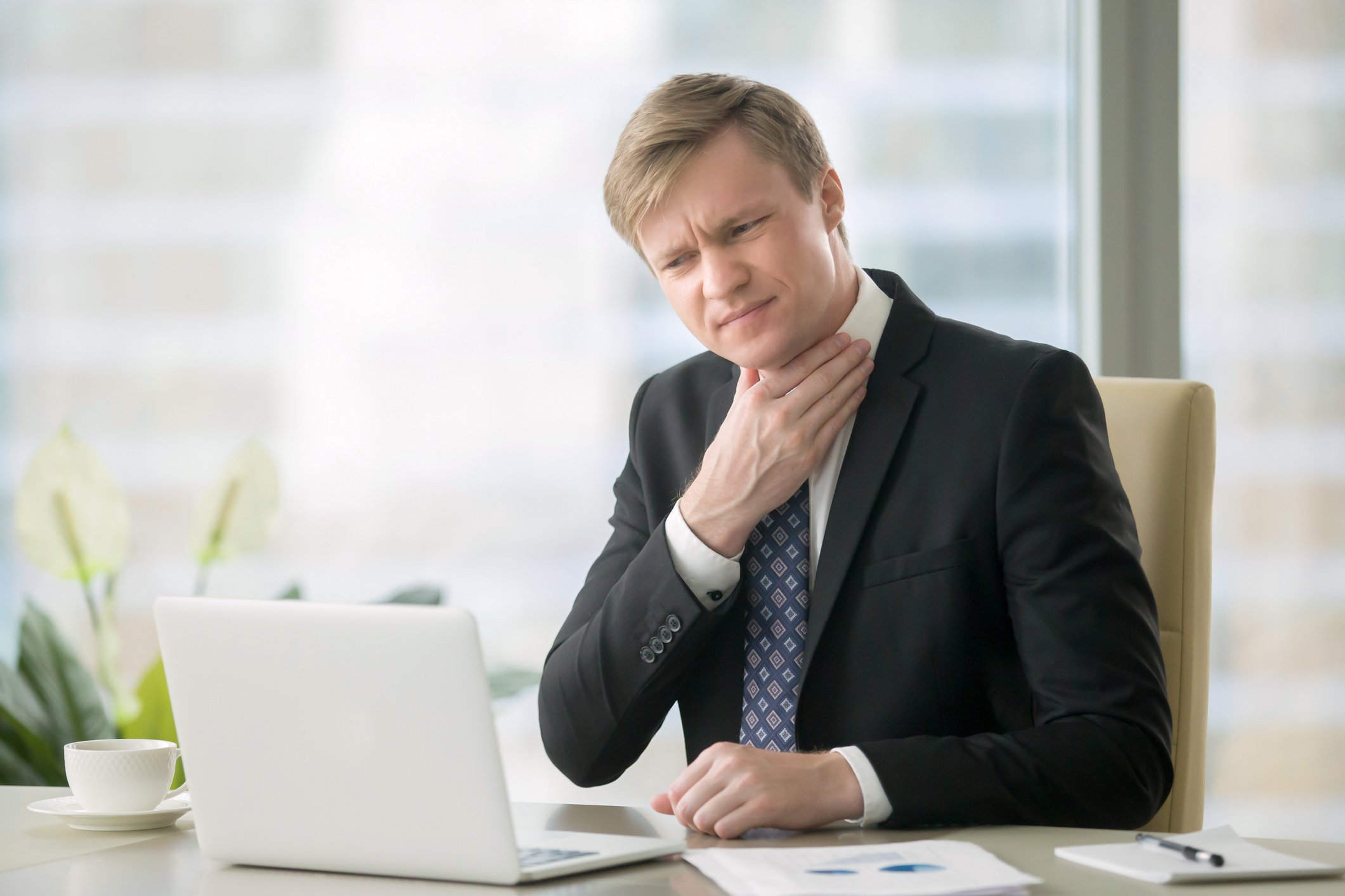 Man in suit at desk holding his throat.