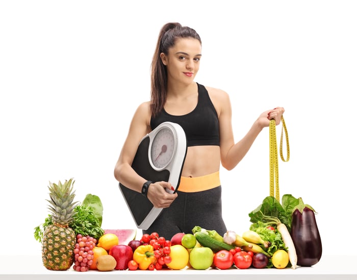 Woman holding scale and tape measure in front of a spread of fruit and vegetables