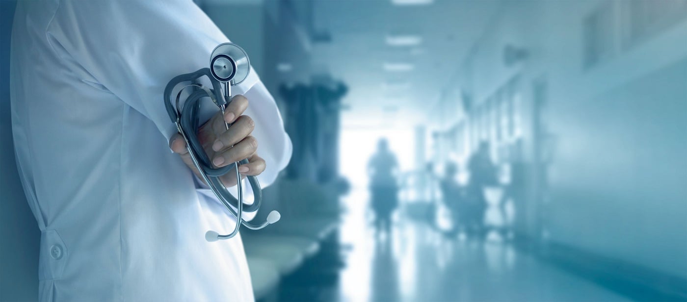 Person in white coat holding stethoscope in a hallway with various people in it.