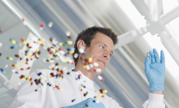Person wearing blue gloves holding a pill, with lots of pills on a glass table.