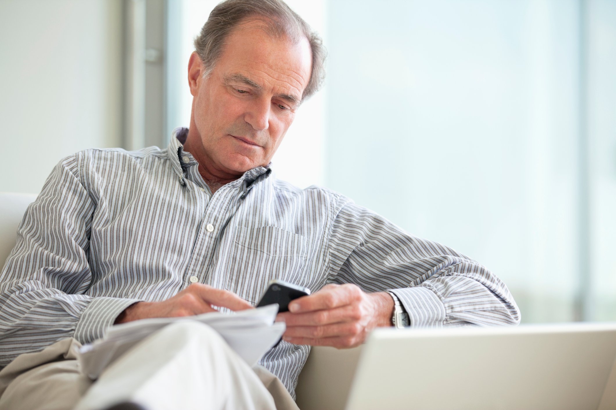 A middle-aged man sits on a sofa, looking at a phone, with a collection of papers on his knee.