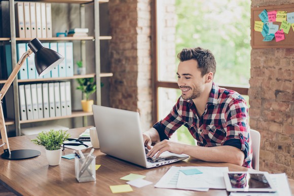 A smiling man sits in an office, typing on a laptop.