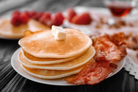 Four pancakes with a pat of butter on top, sharing a plate with some pieces of bacon