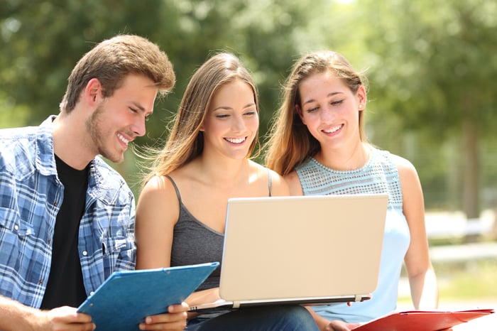 Three students smiling over a shared laptop in the sun