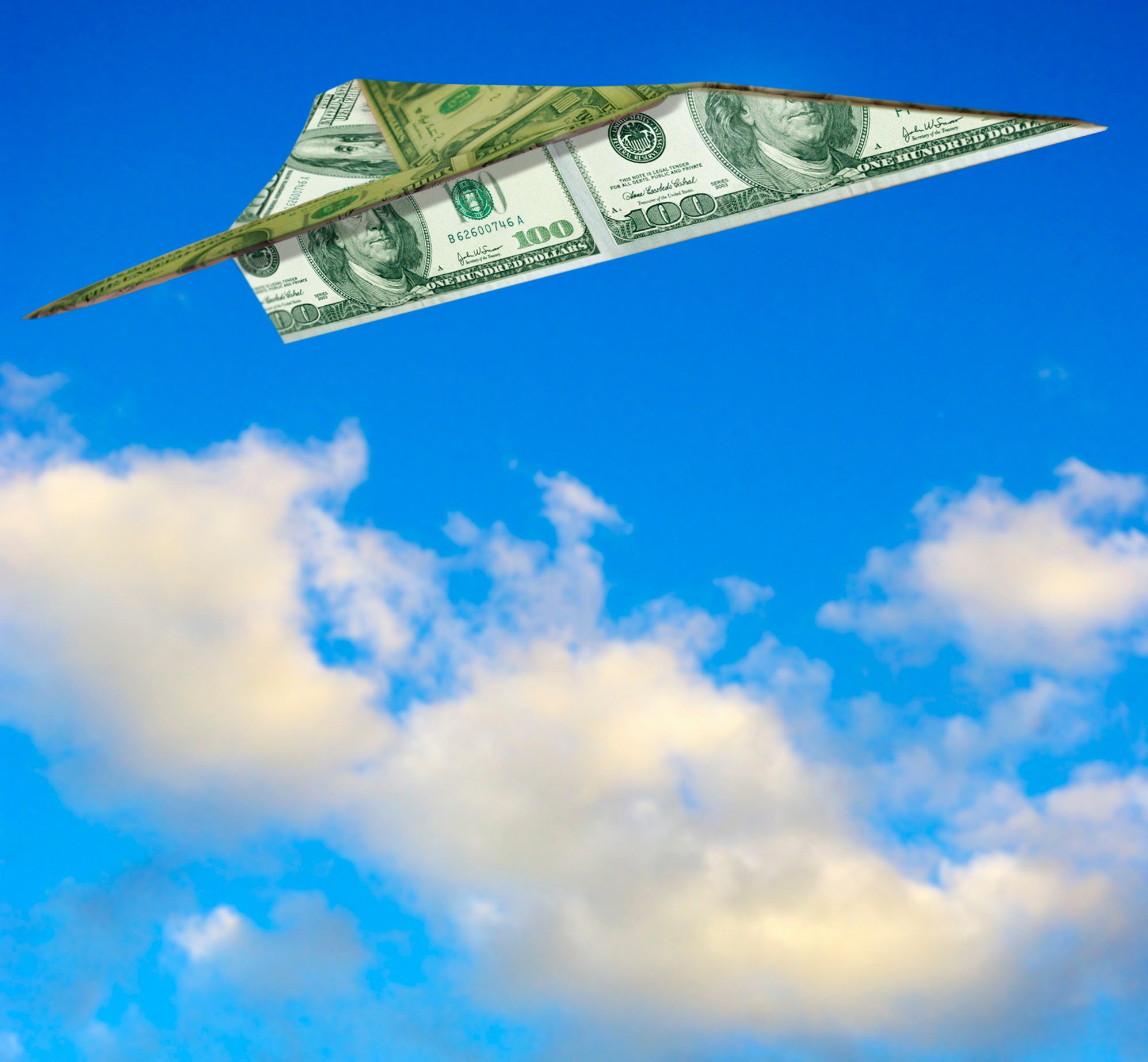 A paper airplane folded out of a $100 bill is flying against a blue sky.