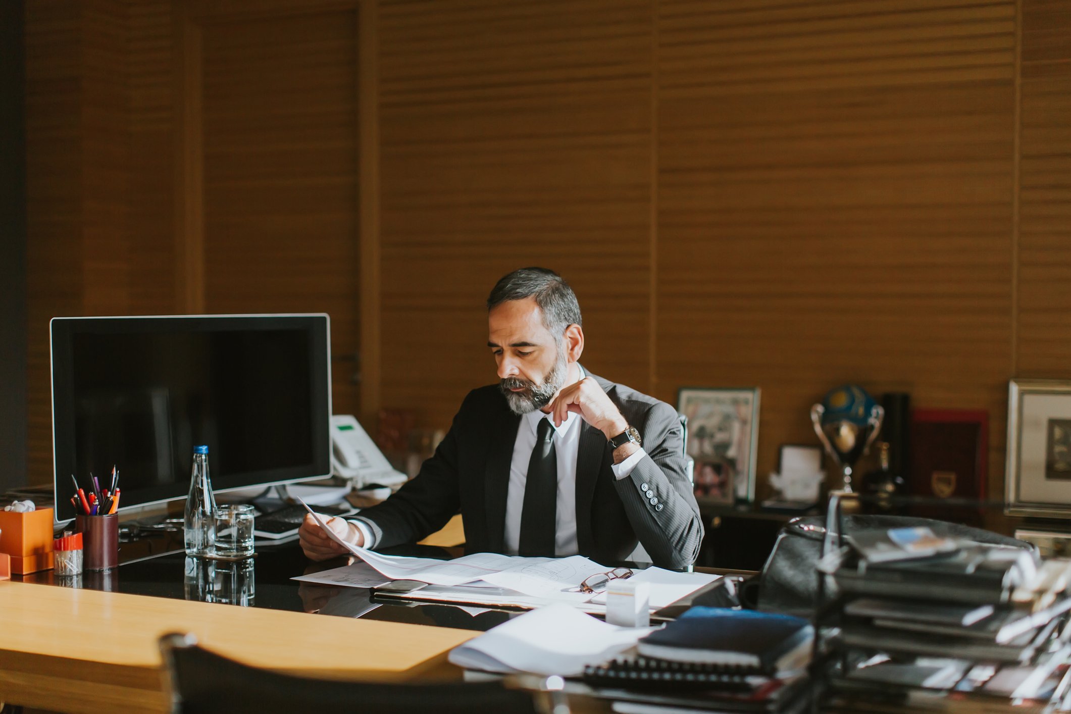 A middle-aged man sits at a desk in an office setting, as he looks thoughtfully at a paper in his hand.