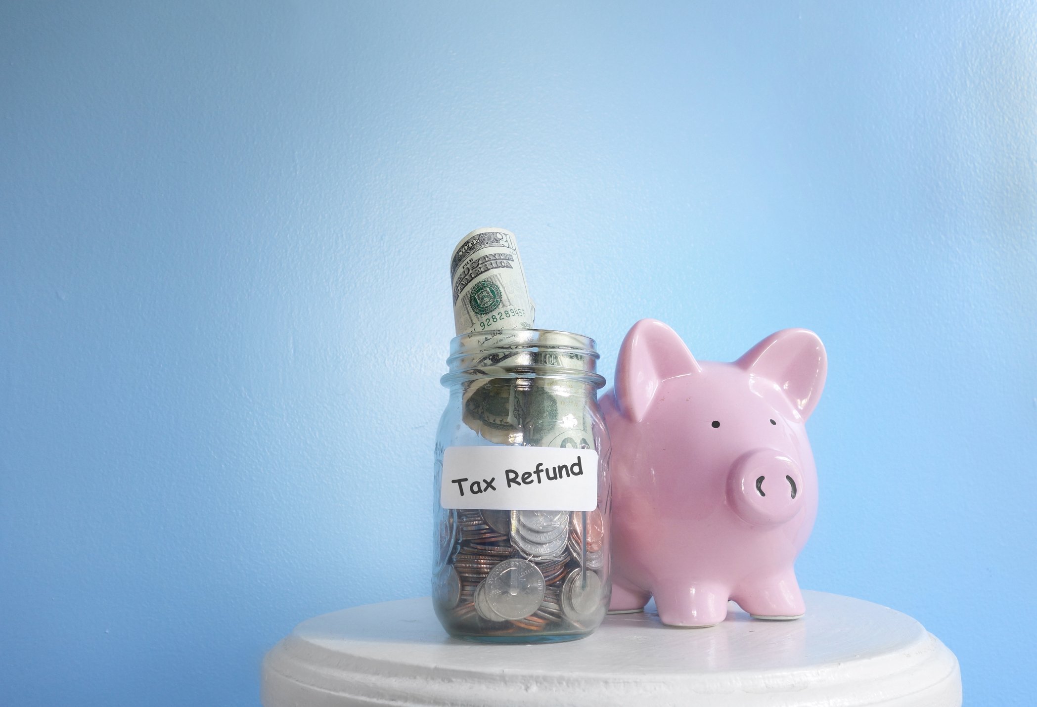 Glass jar with coins and rolled-up bills labeled tax refund next to a pink piggy bank