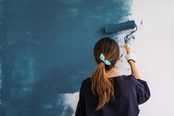 A woman painting a white wall in blue paint with a paint roller.