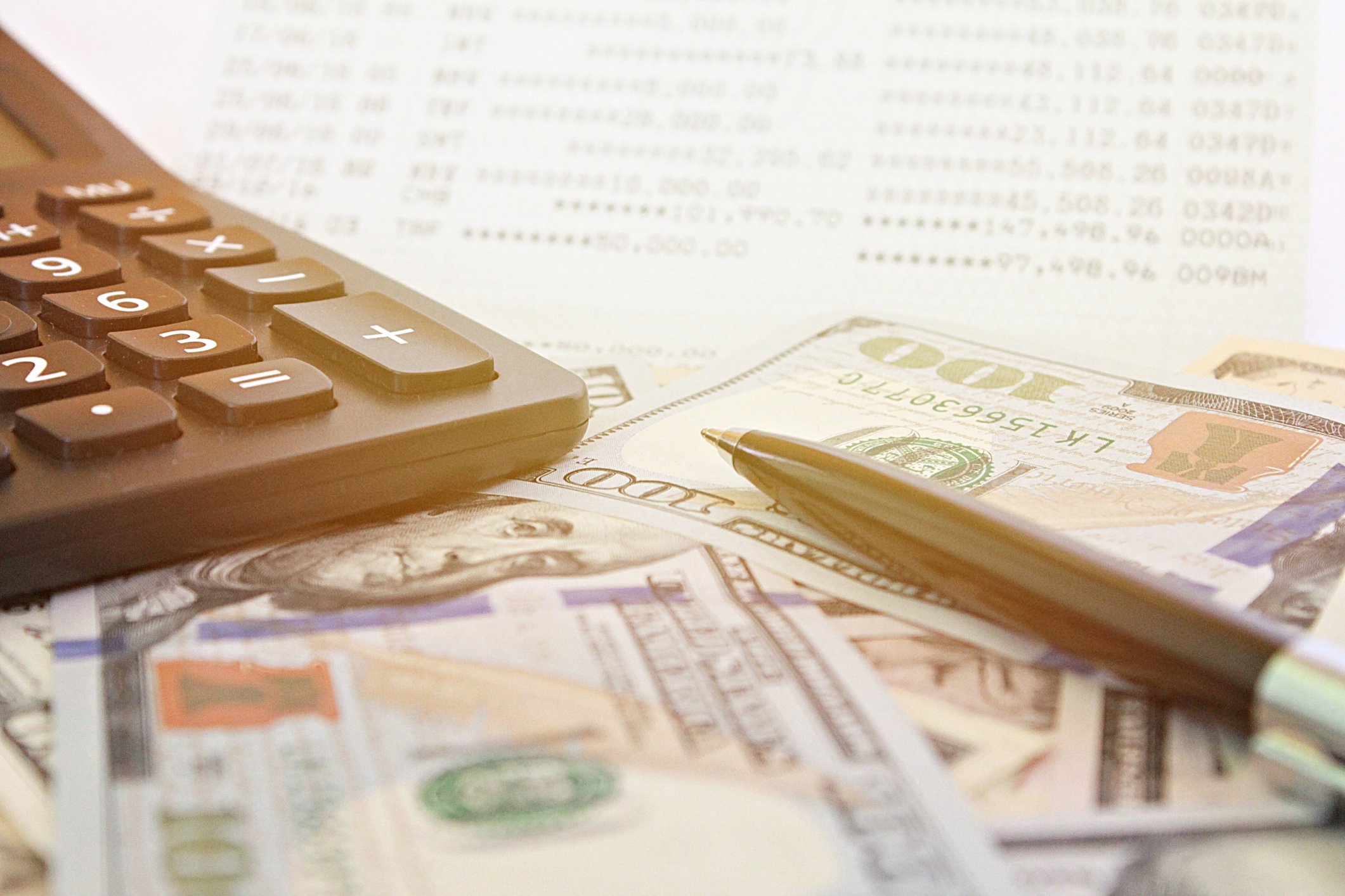 A calculator and pen on top of a pile of U.S. hundred-dollar bills.