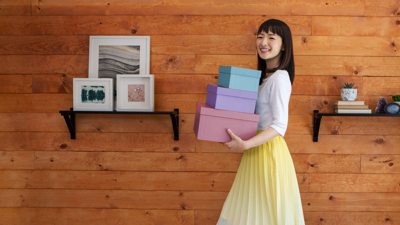 Marie Kondo holding three boxes standing in front of a wall with shelves of pictures and books.