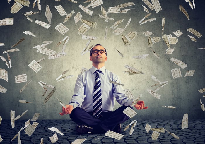 A man sitting in a yoga pose as $100 bills fall from the sky around him.