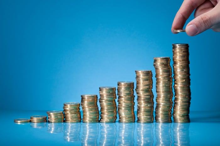 A hand placing a coin on the top of the highest stack of coins in a row of progressively tall stacks of coins.