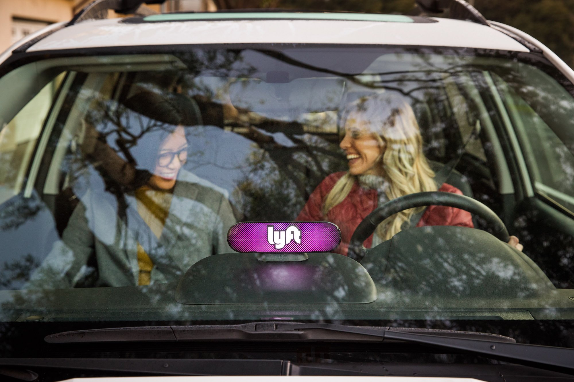 Two smiling women sitting in the front seat of a car with the Lyft logo mounted on the dashboard