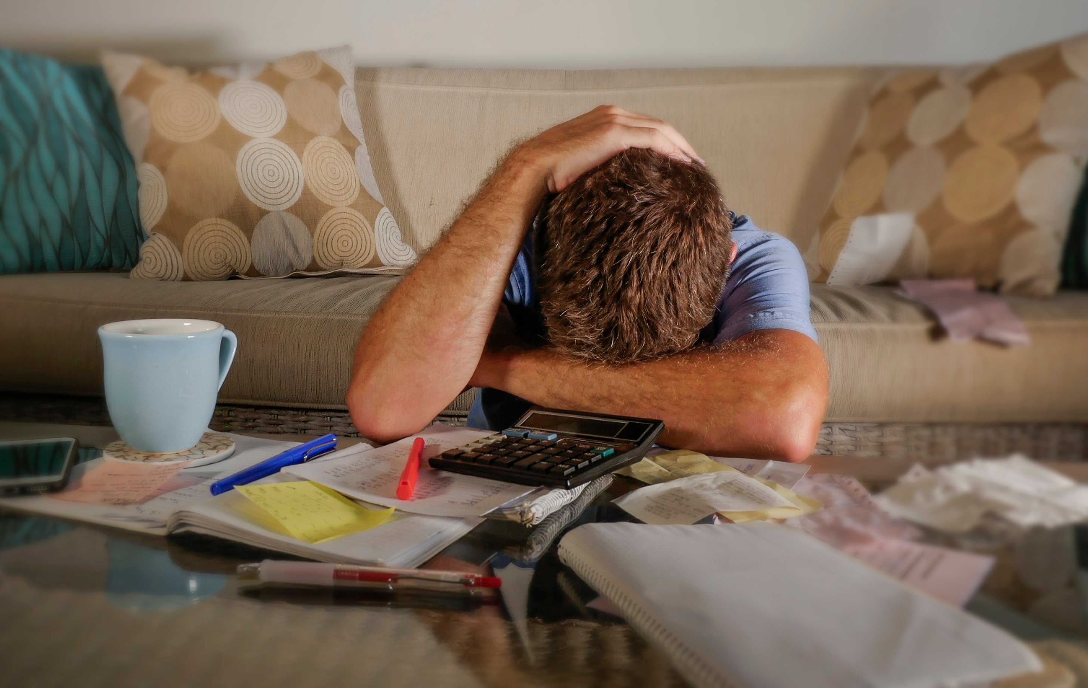 Man with face down on table with papers, pens, post-its, calculator, and cup scattered about.