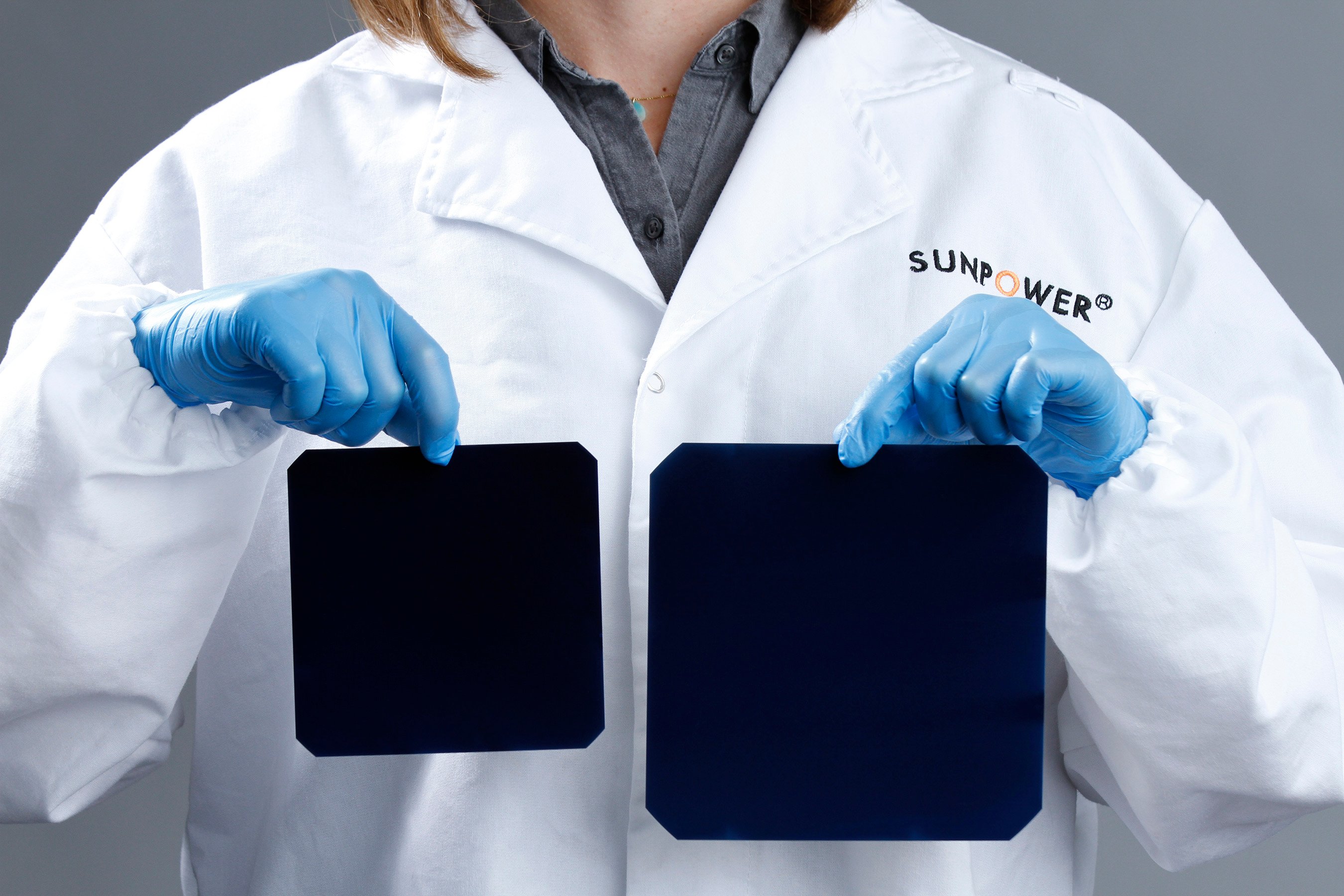 A technician in a lab coat and gloves holding up an A-Series solar cell in one hand and an X-Series in the other