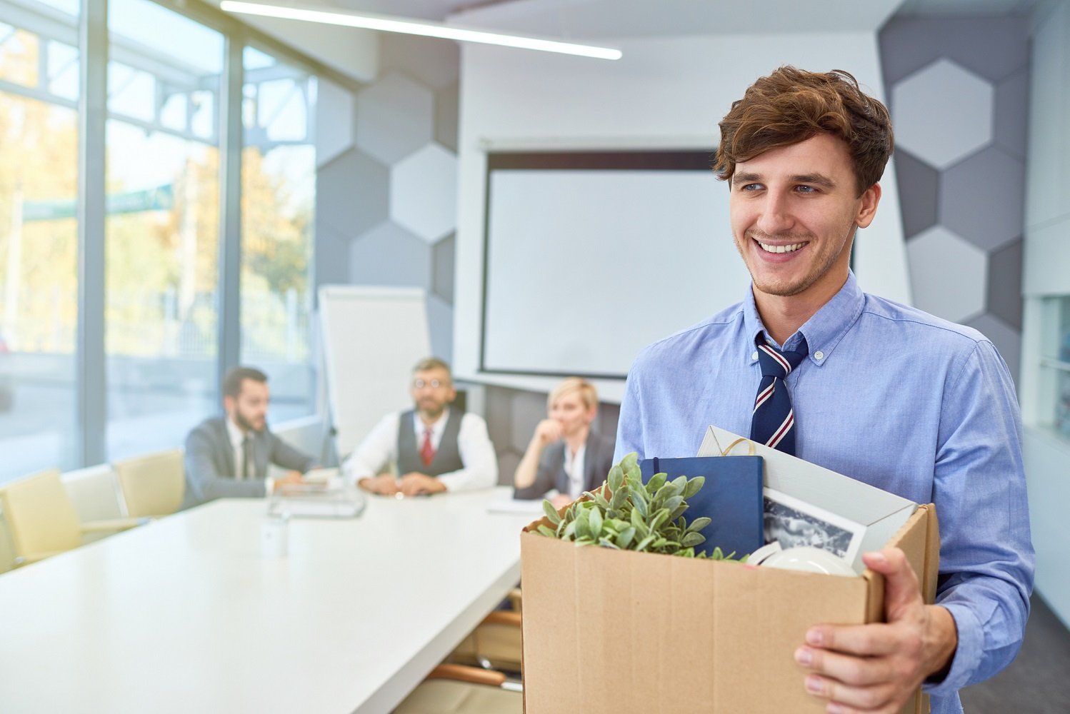 A young man in an office conference room smiles as he carries his office belongings in a box, preparing to walk out.