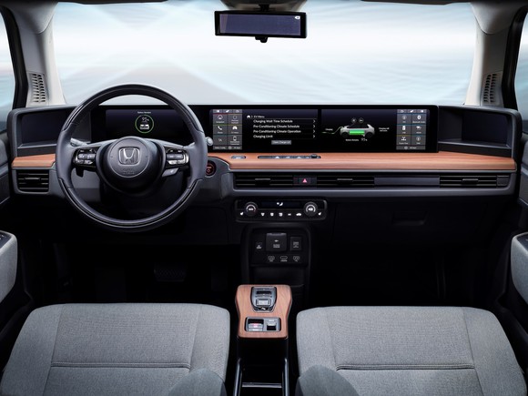 A view of the front seats and dashboard of the Honda E Prototype, showing several touchscreens, reddish wood trim, and typical Honda cloth seats and steering wheel.