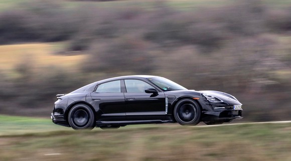 A black Porsche Taycan, a low-slung battery-electric sports sedan, partially covered in fabric to conceal its lines, is shown testing on a country road near Porsche's research center in Weissach, Germany. 