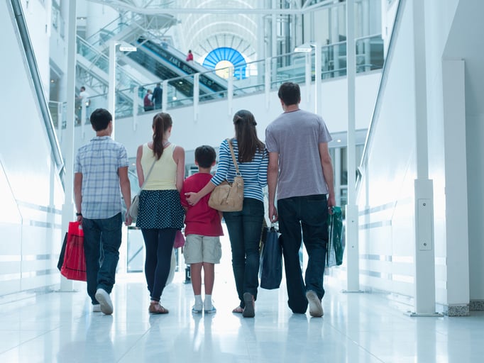 The back view of a man, woman, and three children, all carrying bags, as they walk in a shopping mall.