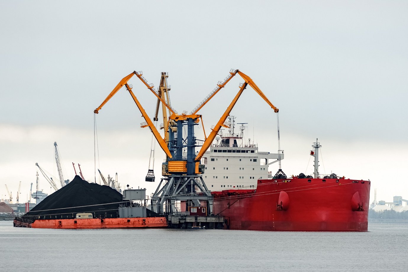 Red ship taking on cargo at a port, with large crane arms helping to load.