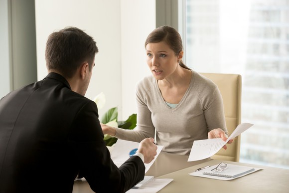 In an office setting, a woman holding papers sits looking frustrated across a desk from a man in a suit.