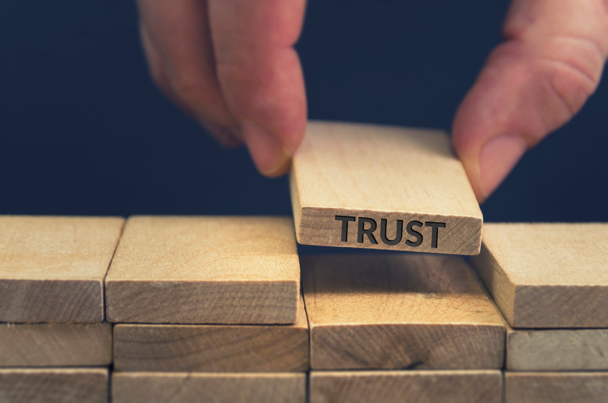 The word "trust" etched into a wooden block that's being placed into a stack of wooden blocks.