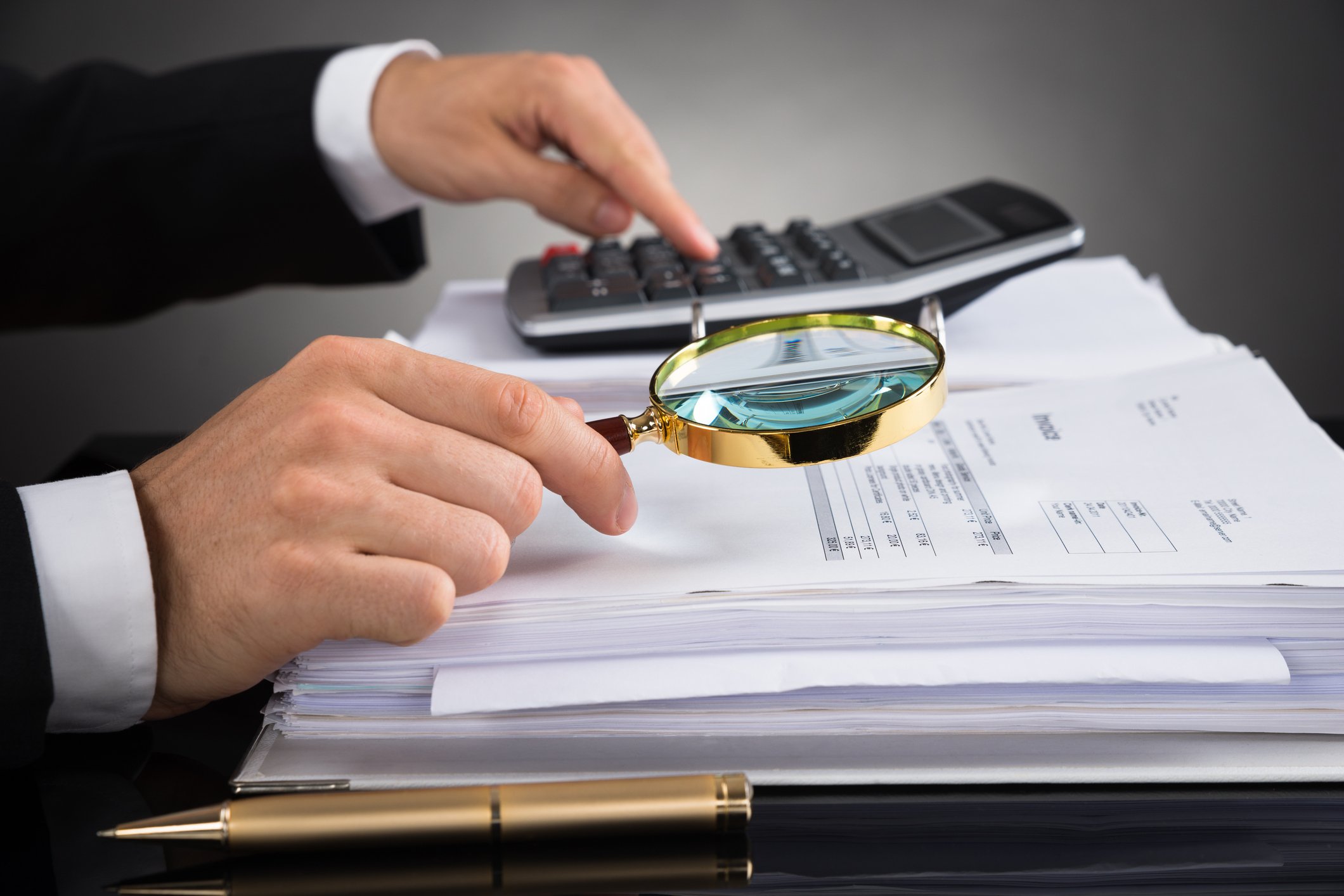 Man typing on calculator and holding magnifying glass to a pile of papers