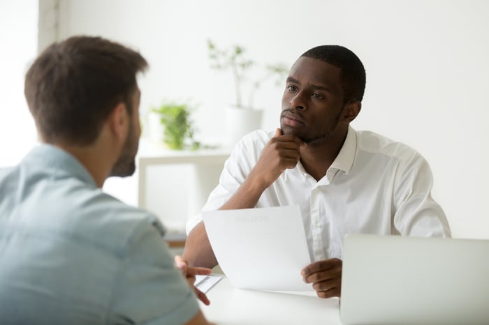 Man holding sheet of paper sitting across from another man with hand on chin, as if listening intently