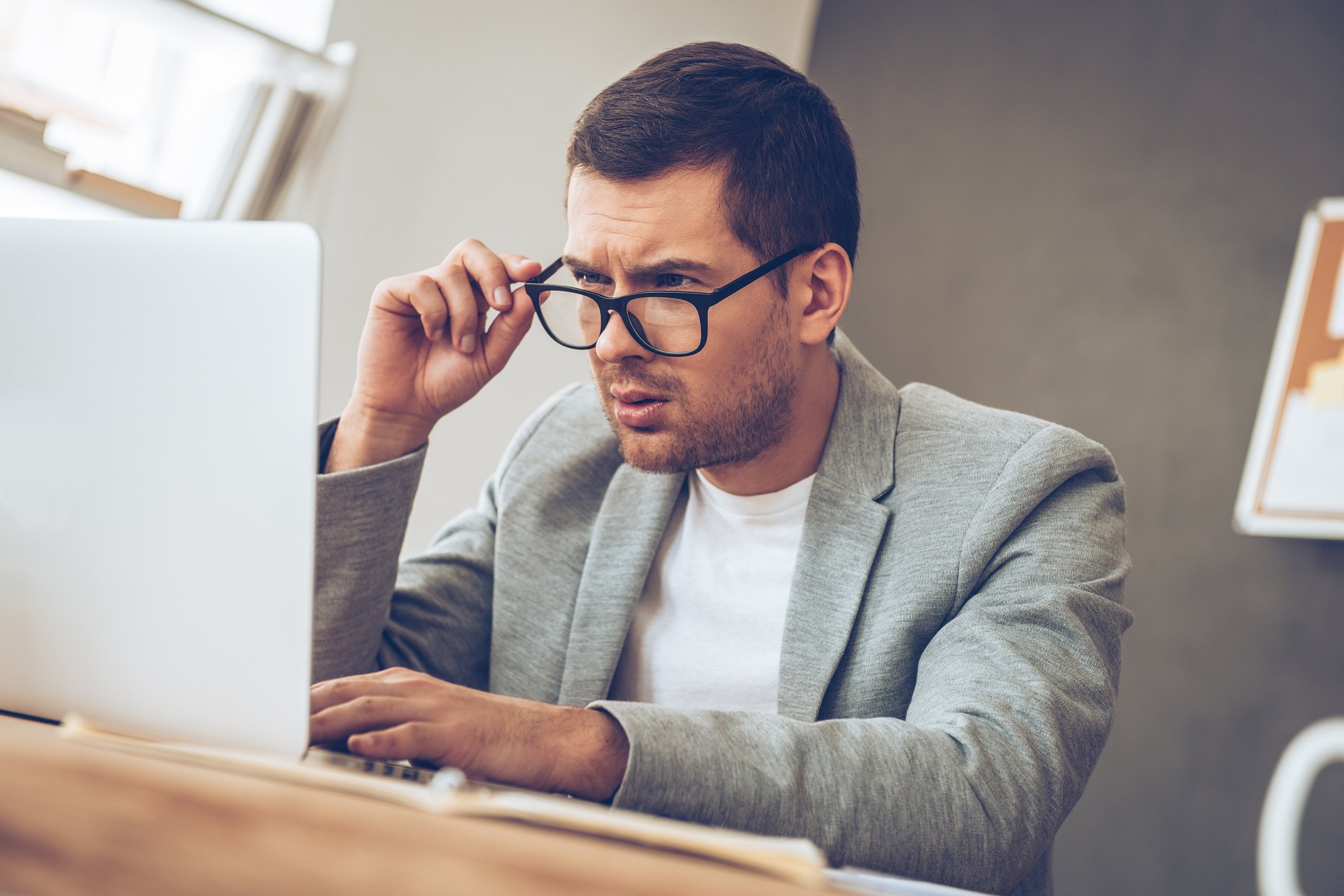 Man in grey jacket and white tee shirt at computer adjusting his glasses.