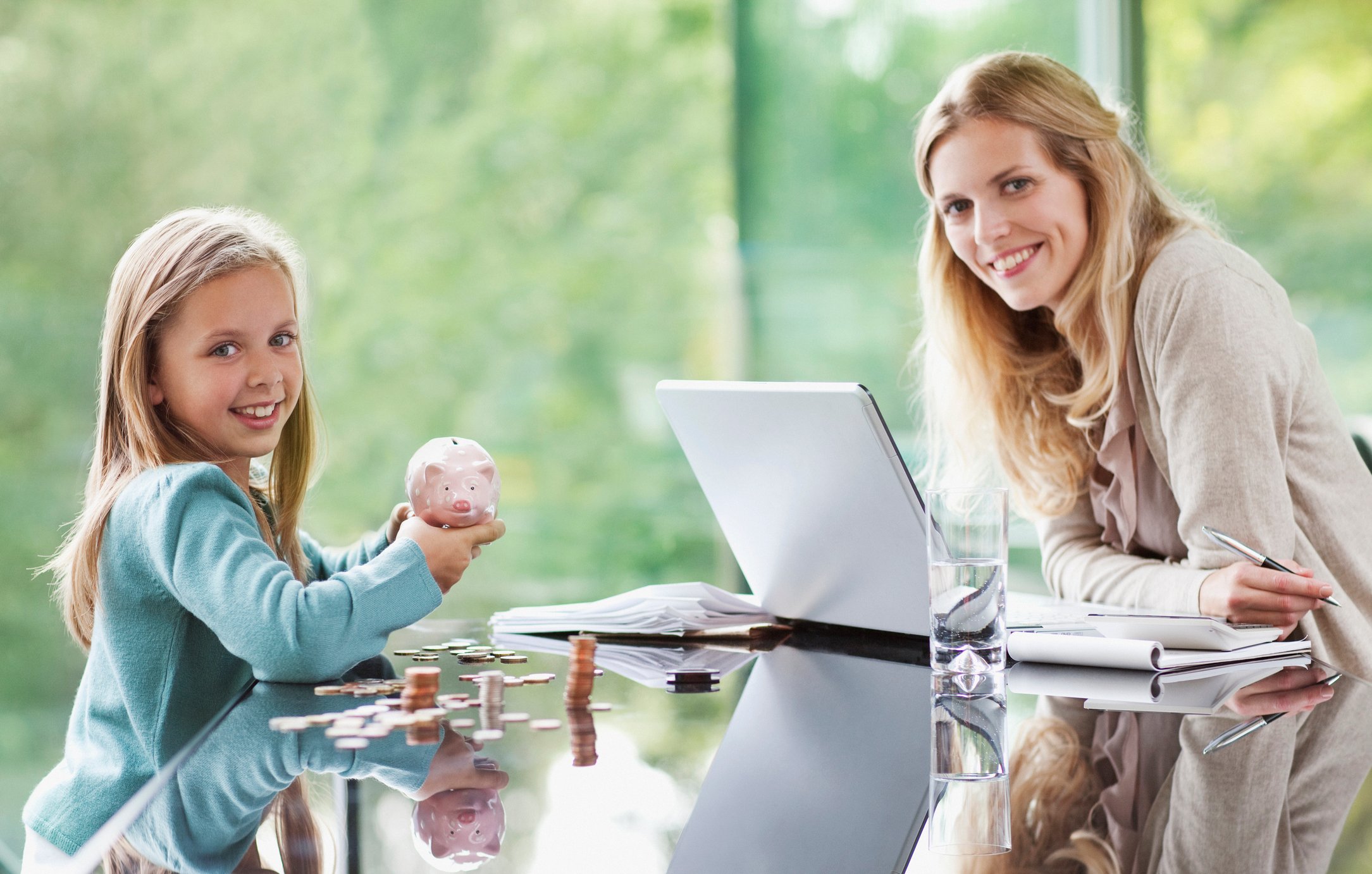 A child and an adult with similar appearance sitting at a glass table. The child holds up a piggy bank.