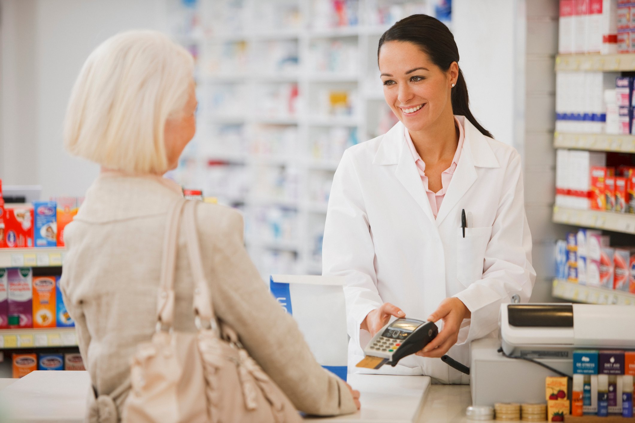 Woman in white coat in a pharmacy holding credit card reader out to woman across the counter.