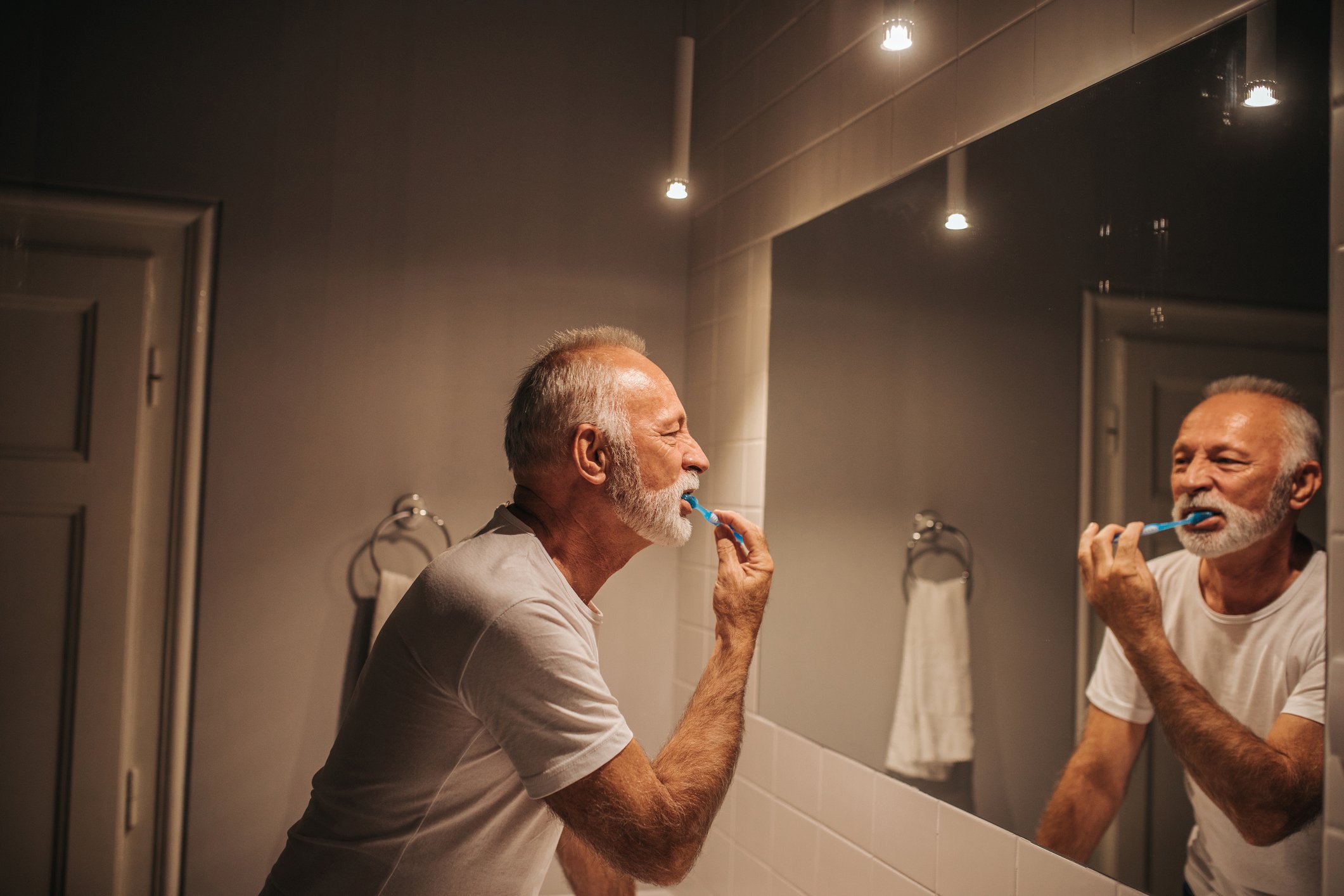 A senior man looks in the bathroom mirror as he brushes his teeth.