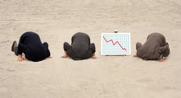 Three businessmen sticking their heads in the sand as a chart next to  them shows a red down arrow. 