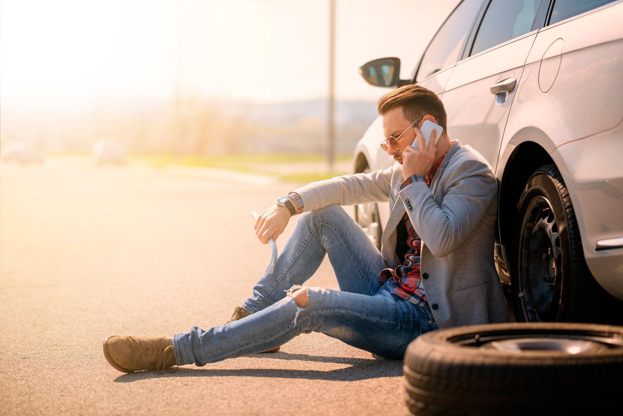 Man sitting against car on the side of the road, talking on a smartphone