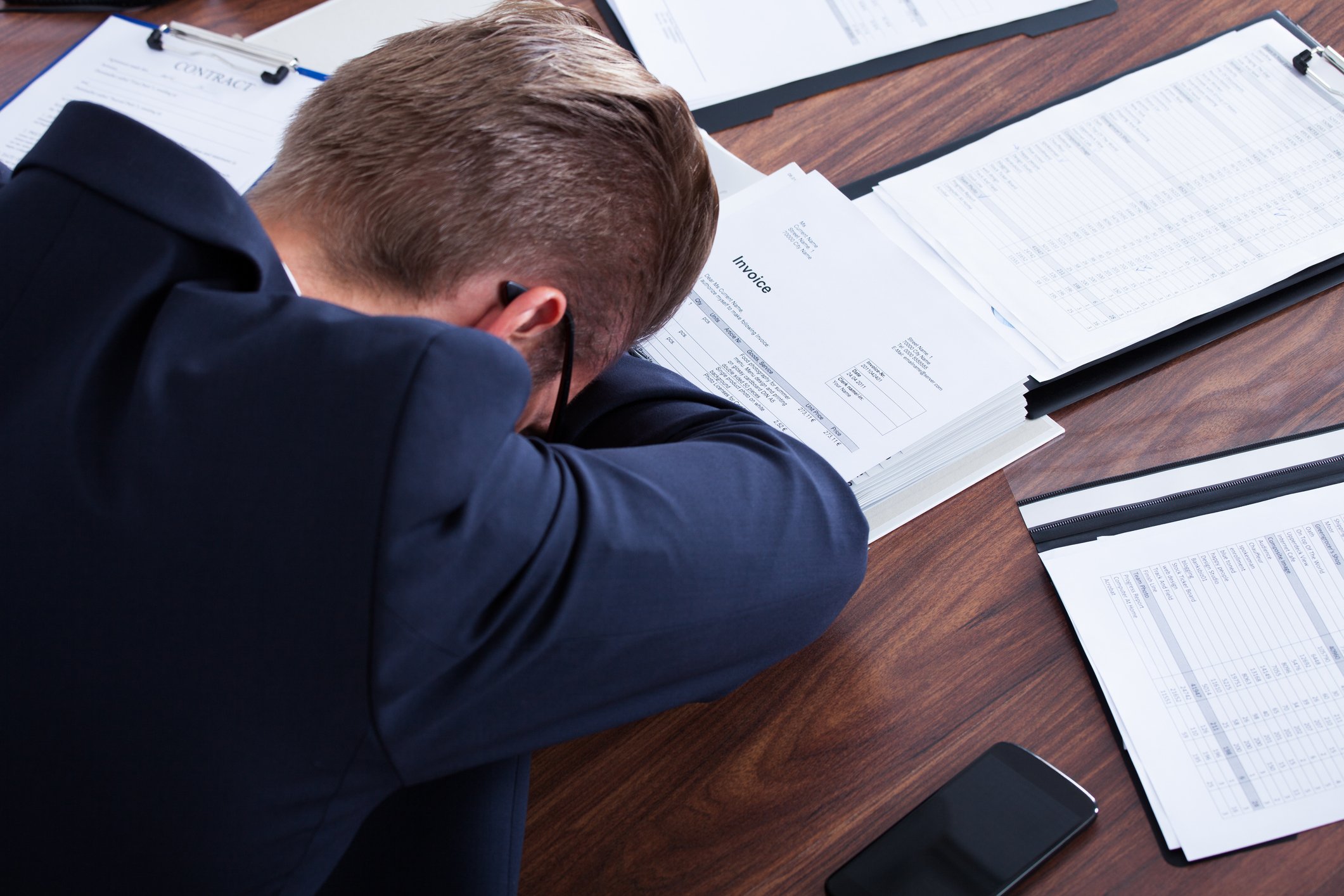 A man in business attire puts his head down on a desk covered with documents.