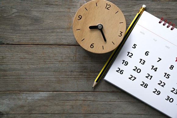 A brown clock next to a pencil and a calendar on top of a table.