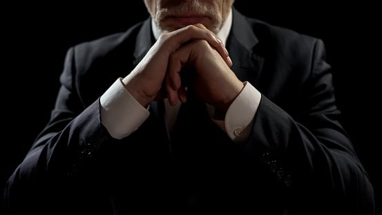 A man in a suit viewed from the neck down rests his chin on folded hands which rest on a table below. 