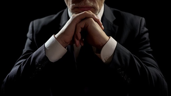 A man in a suit viewed from the neck down rests his chin on folded hands which rest on a table below. 