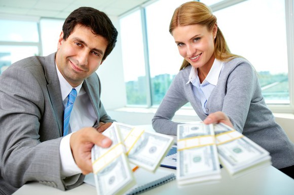 A man and a woman handing stacks of $100 bills to the viewer.
