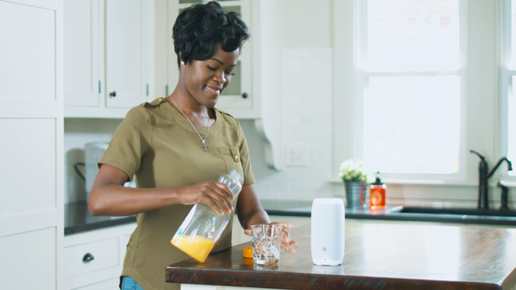 A woman pouring orange juice into a glass that's on a kitchen island with a Google Home speaker on it.