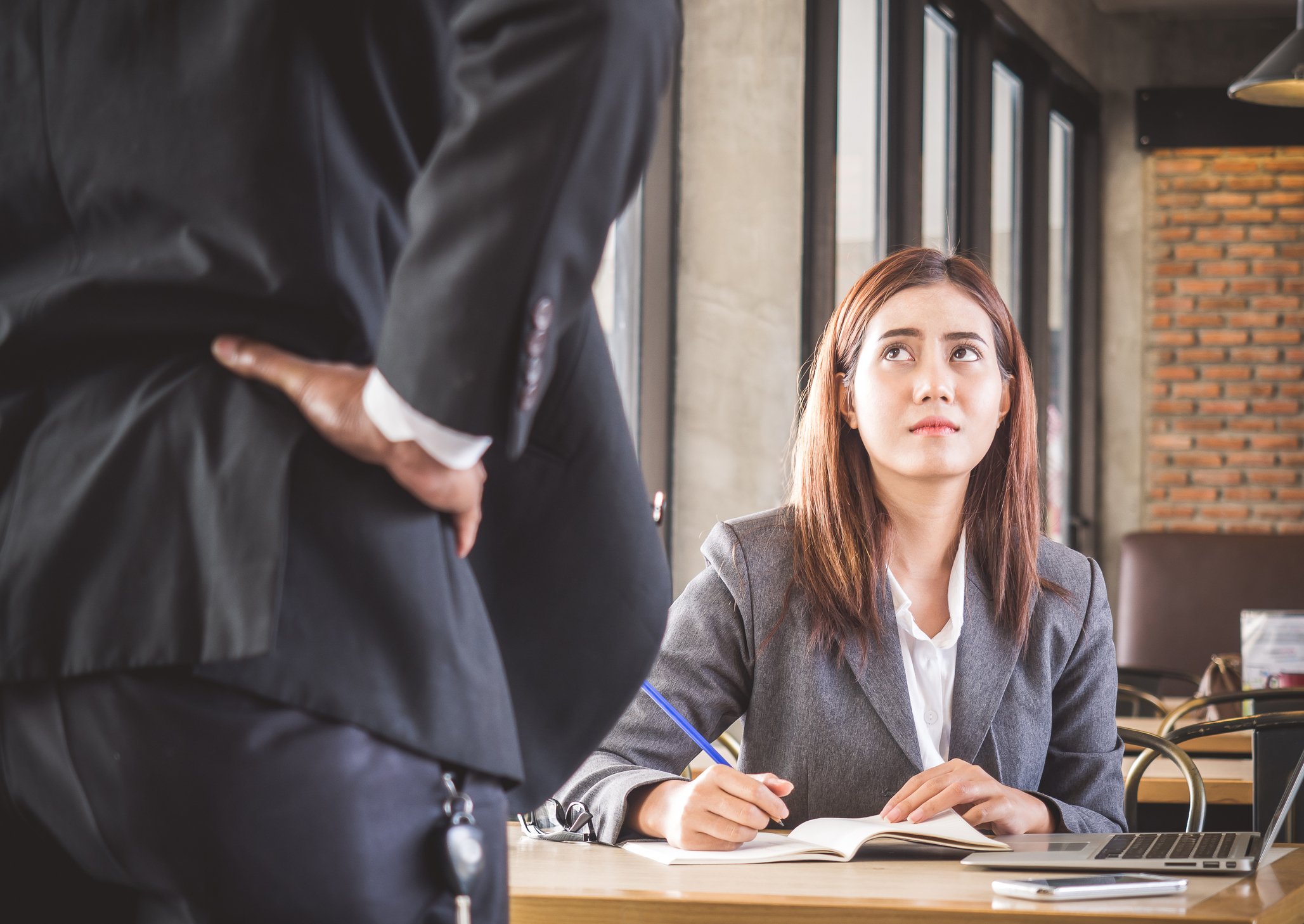 Woman in business suit seated at desk with annoyed expression looks up at man in suit who has his hands on his hips.