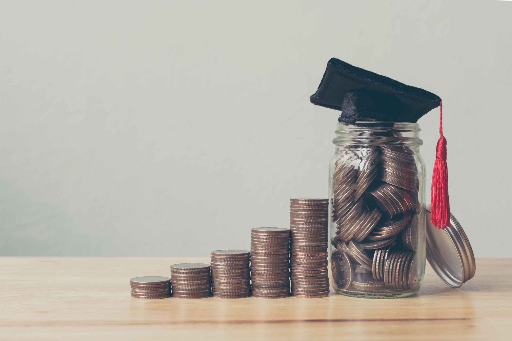 Progressively higher stacks of coins next to a jar filled with coins with a graduation cap on top of it. 
