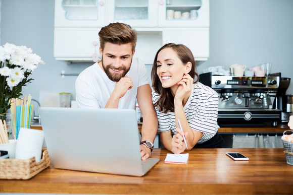 Young man and woman behind the counter of what looks like an eatery or coffee shop and looking at a laptop screen.
