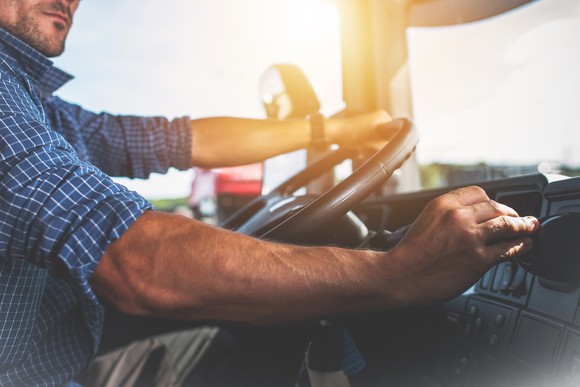 A semi-truck driver inside his cab with his left hand on the steering wheel.