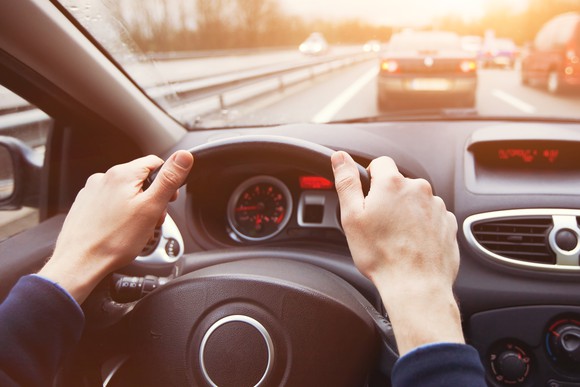 Man with hands on car's steering wheel; cars ahead on the road are visible from windshield
