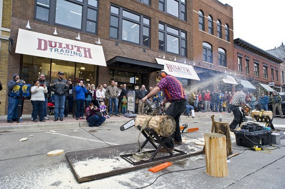 Person in flannel shirt cutting log on a street in front of a Duluth Trading Company store location.