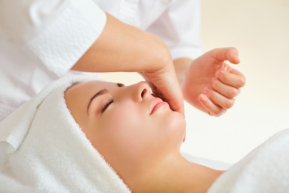 Close-up of a woman's face and head, with her hair covered with white terry cloth, as she gets a facial massage. 