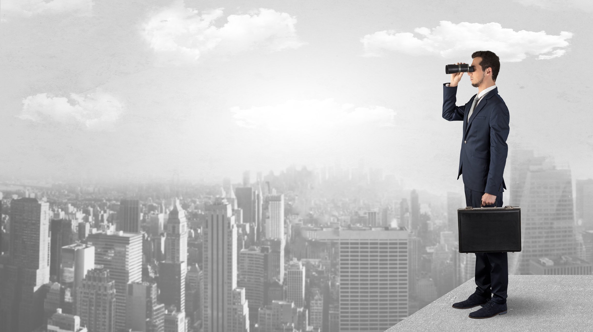 Guy in a suit standing on a rooftop and looking ahead with binoculars.